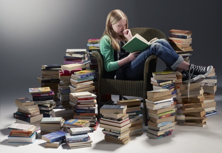 The girl reads a book in a chair surrounded by piles of other books